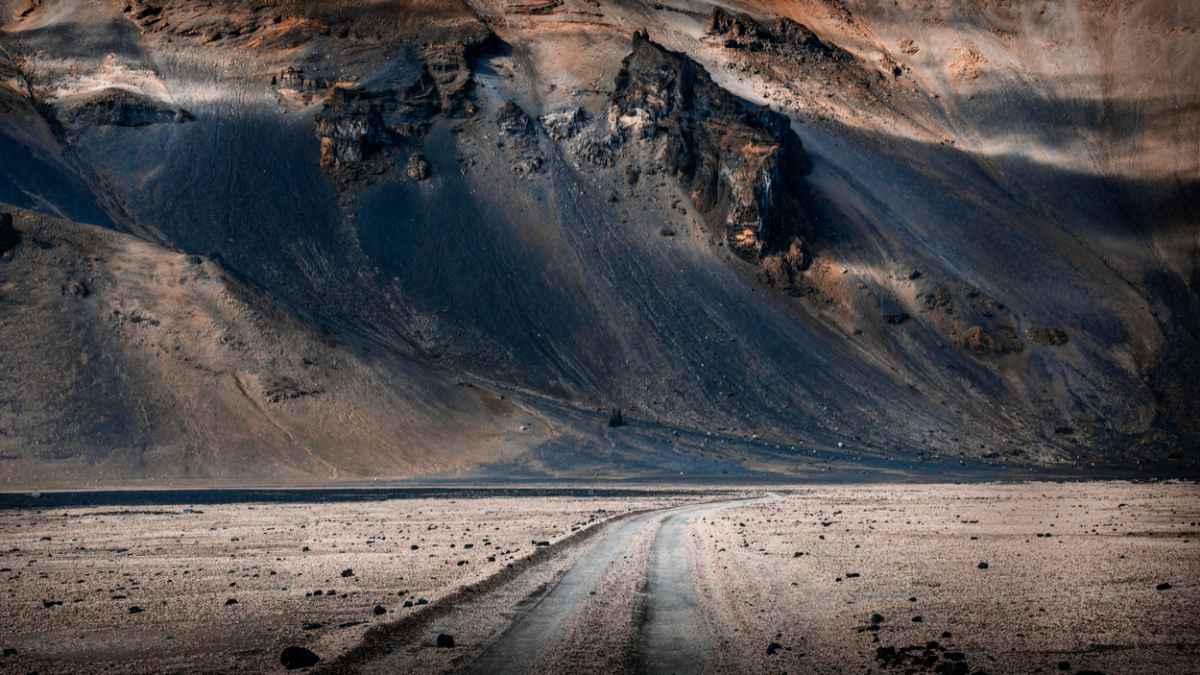Desolate dirt road in Iceland leads to rugged mountains under overcast sky, with dark rocky slopes and scattered stones creating a barren, remote scene.