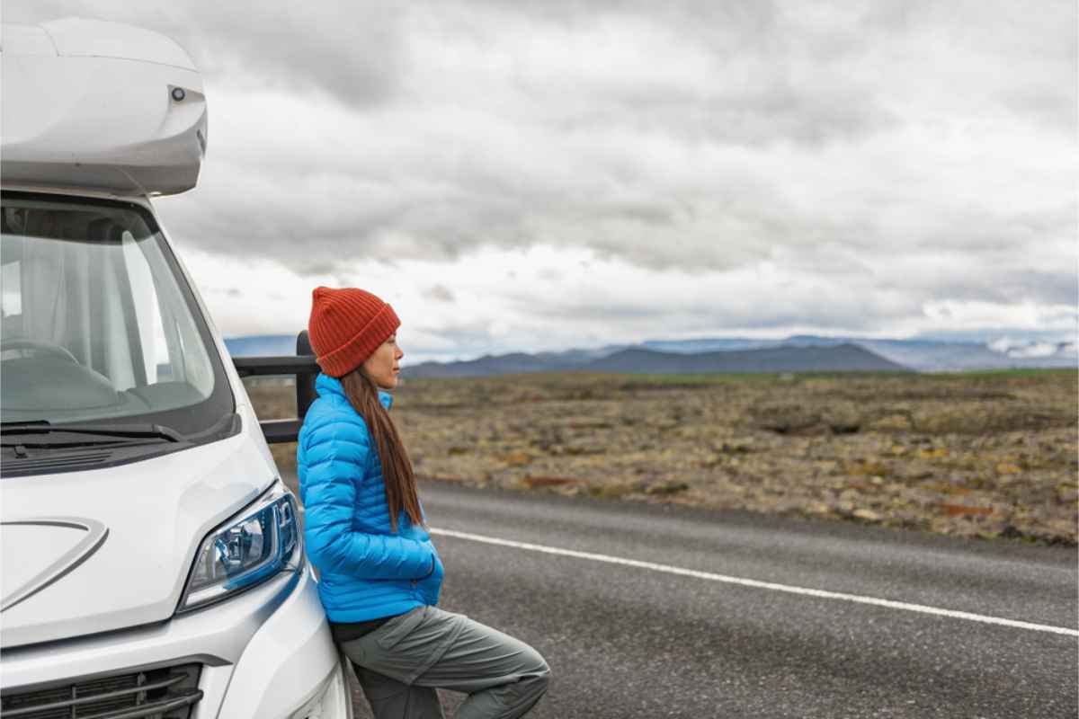Woman in a blue jacket and red beanie leans on a white RV, gazing at a vast, cloudy landscape. Overcast sky, peaceful mood.