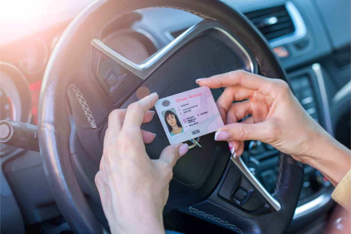 Hands holding a driver’s license in front of a car steering wheel. The license is pink with a photo and text. The car interior is visible.