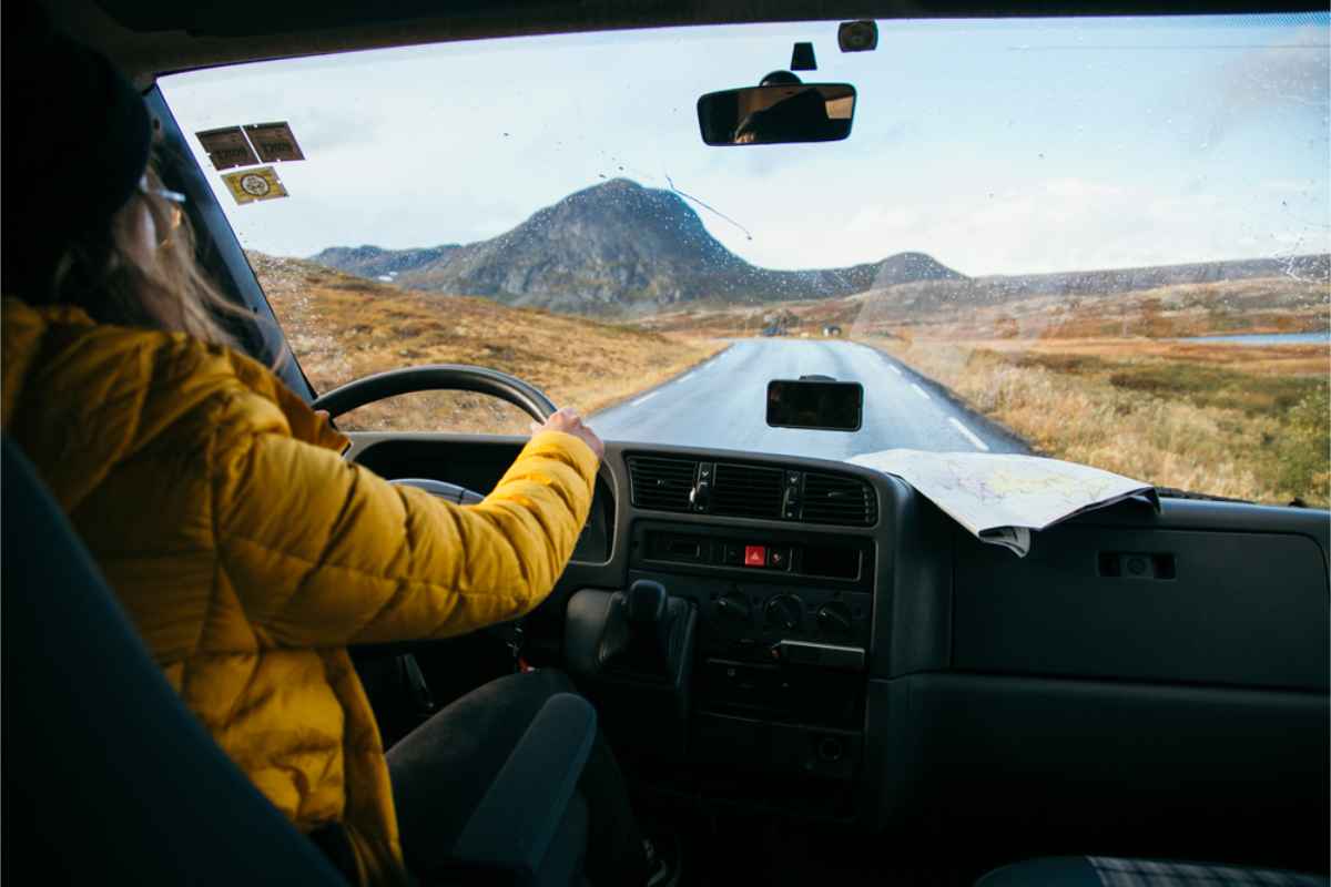 Person in a yellow jacket drives a van on a scenic road in Iceland with mountains. A map rests on the dashboard, creating an adventurous mood.