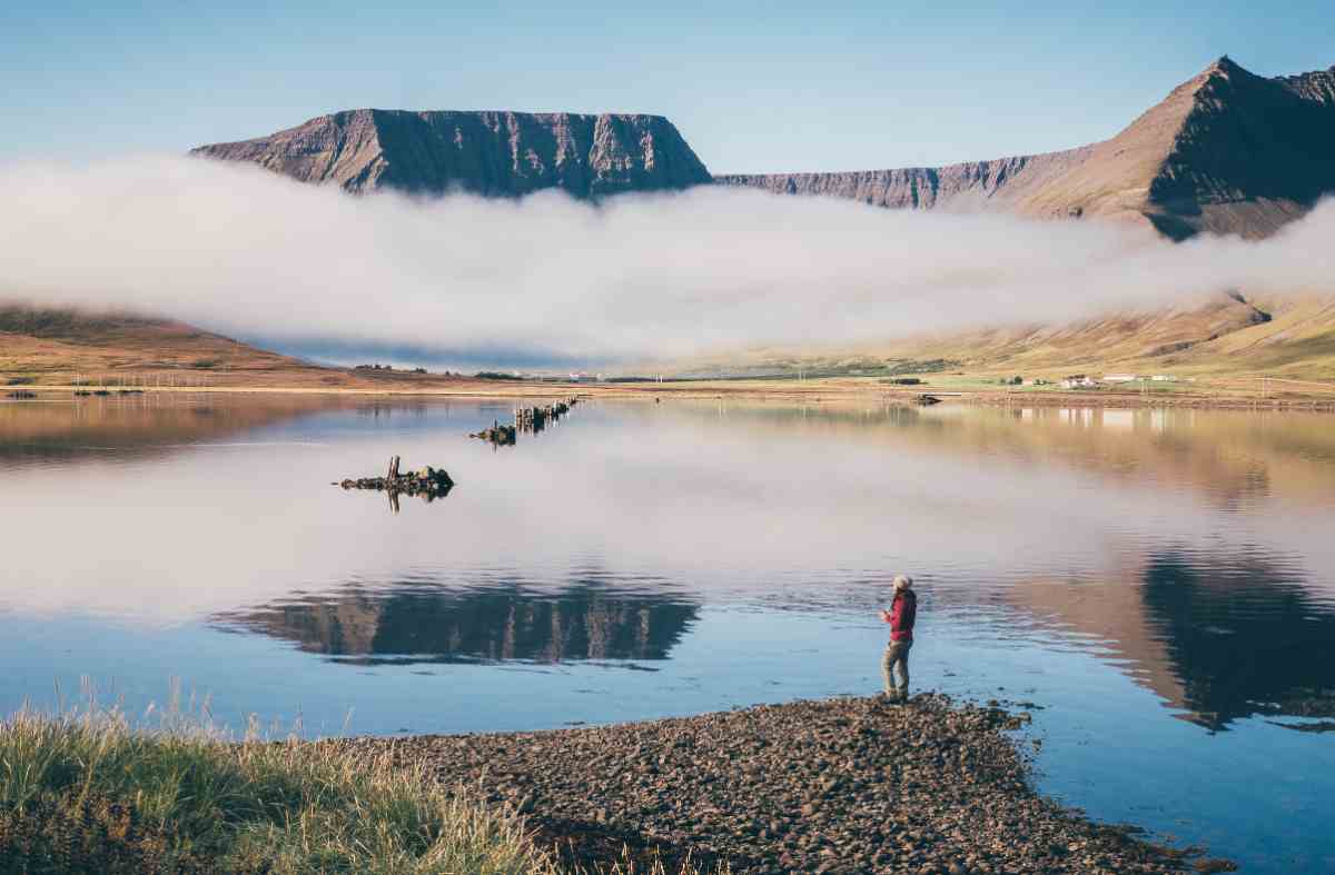 Quiet Westfjords scene with low fog over mountains and a person standing by a mirror-calm fjord.