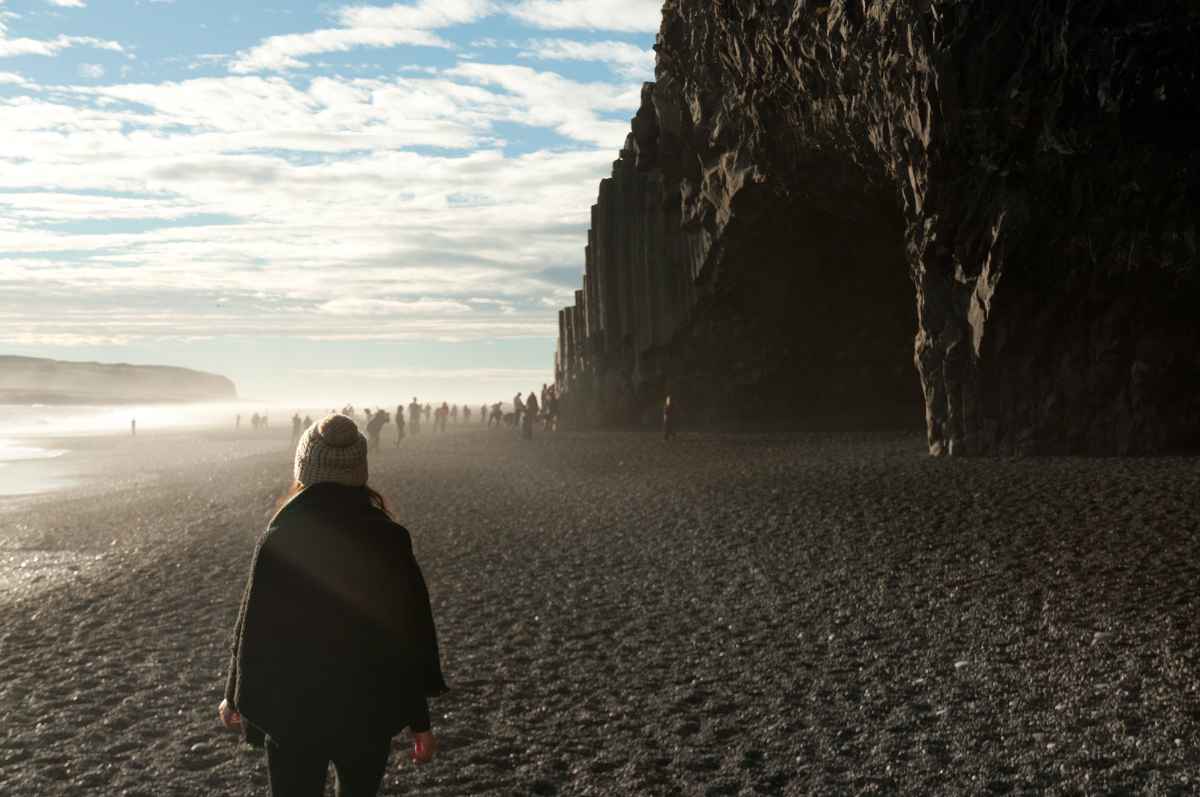 Visitor walking along the black sands at Reynisfjara toward the basalt-column cave on a bright, hazy day.