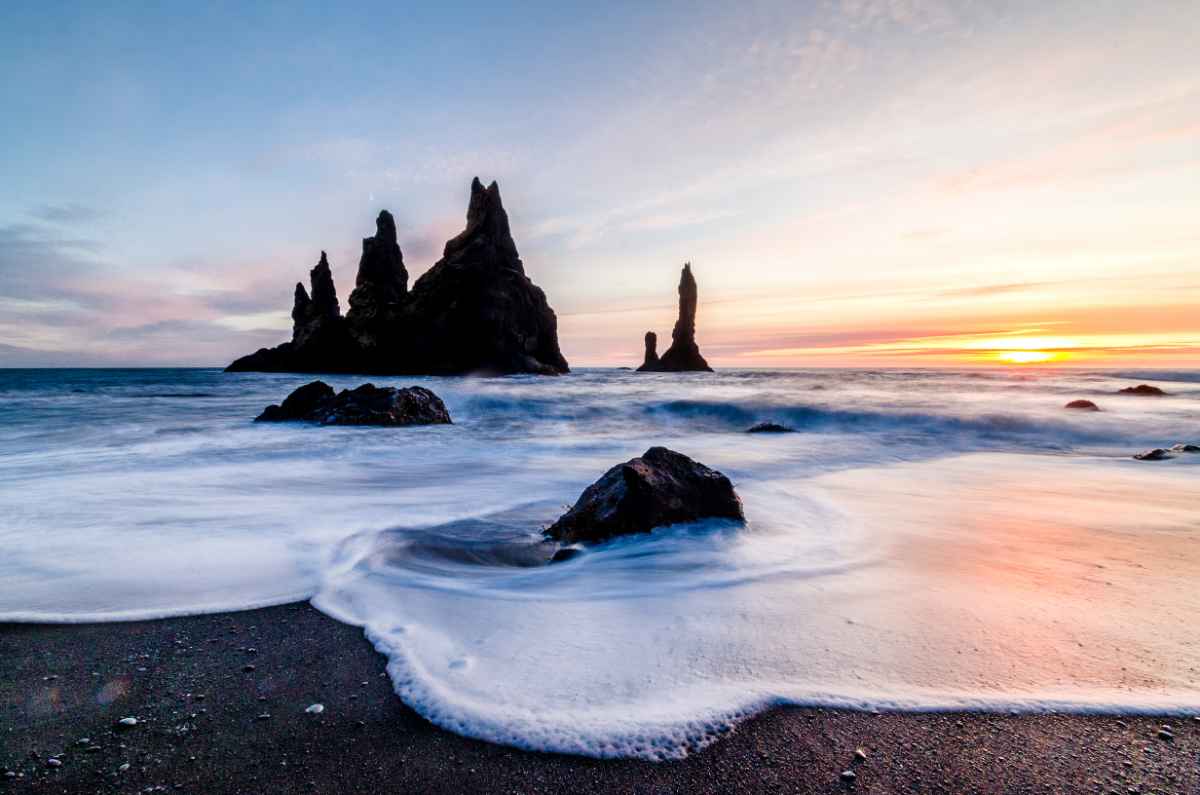Sunset light behind the Reynisdrangar sea stacks, waves swirling over black sand at V&iacute;k.