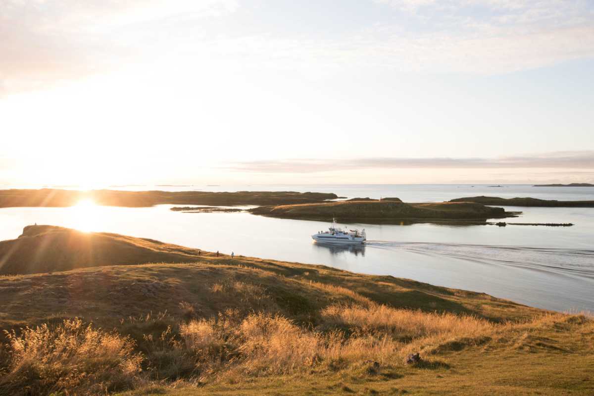 Passenger ferry gliding through sunlit waters among low grassy islets at sunset.