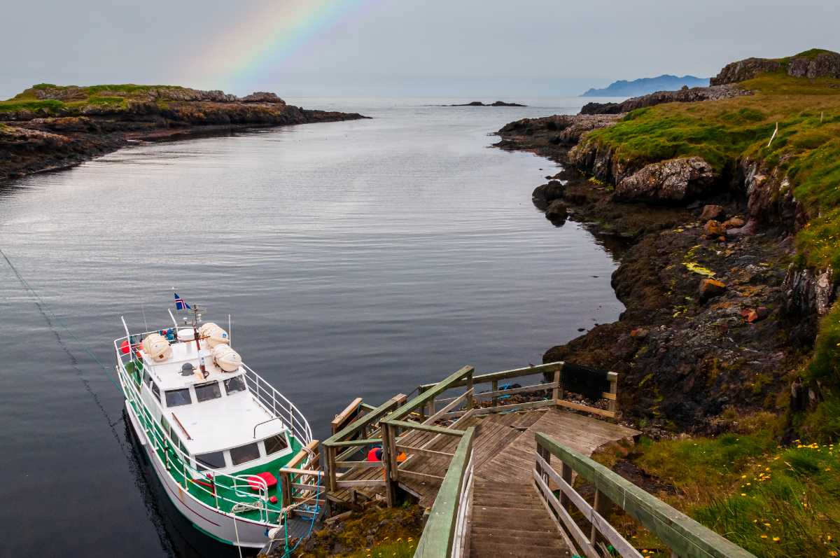 Small passenger boat tied to a wooden dock in a quiet cove, faint rainbow arching overhead.