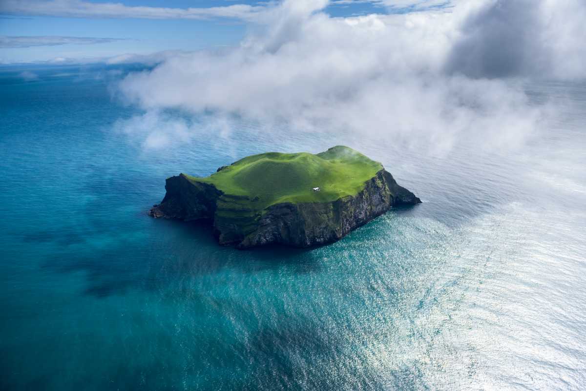 Aerial view of a tiny green island with a lone white house surrounded by turquoise ocean and low clouds.