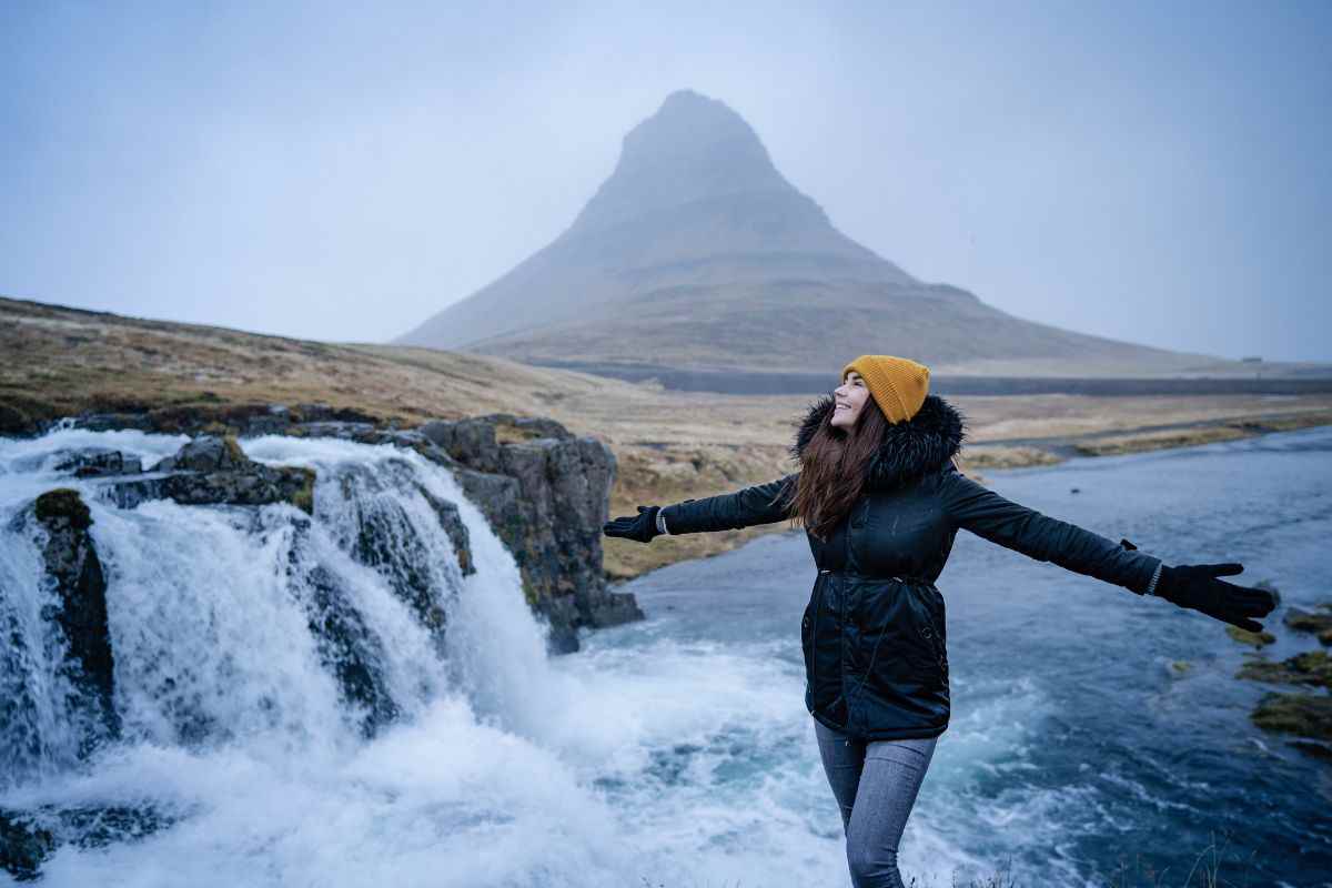 Traveler in a yellow beanie smiling with arms outstretched beside Kirkjufellsfoss, misty winter backdrop of Kirkjufell mountain in Iceland.