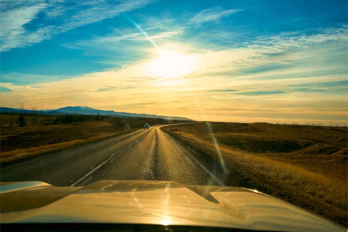 Paved road at sunset seen through a windshield, low sun with lens flare.