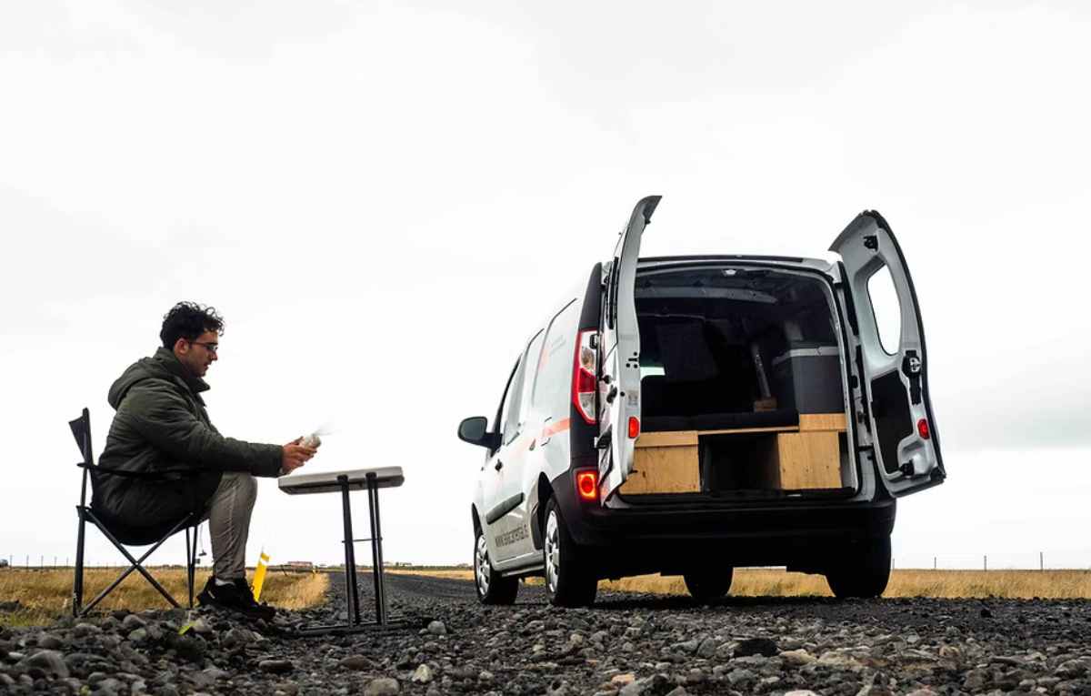 Camper cooking at a folding table beside a small campervan with rear doors open on a gravel roadside.