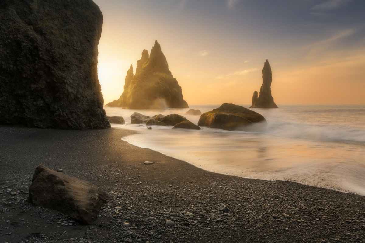 Reynisdrangar sea stacks glowing at sunset above black sand and gentle surf.