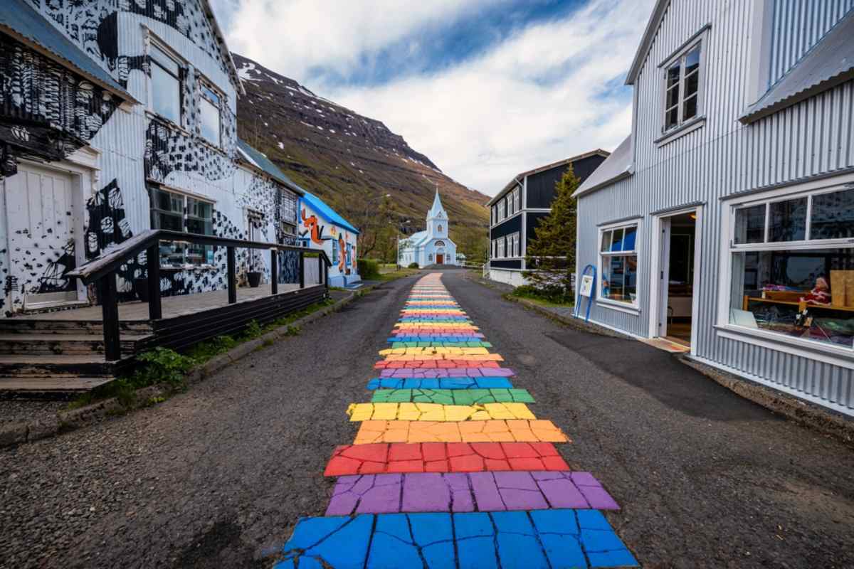 Rainbow-painted street leading to the blue church in Sey&eth;isfj&ouml;r&eth;ur, lined with tin-clad houses.