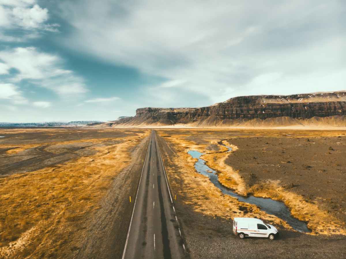 Aerial view of the Ring Road stretching across golden plains, campervan parked by a winding stream and cliffs.