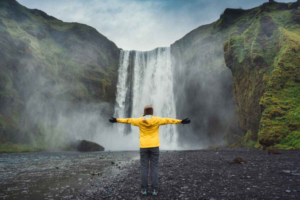 Visitor in a yellow jacket facing Sk&oacute;gafoss waterfall, arms outstretched in mist at the base.