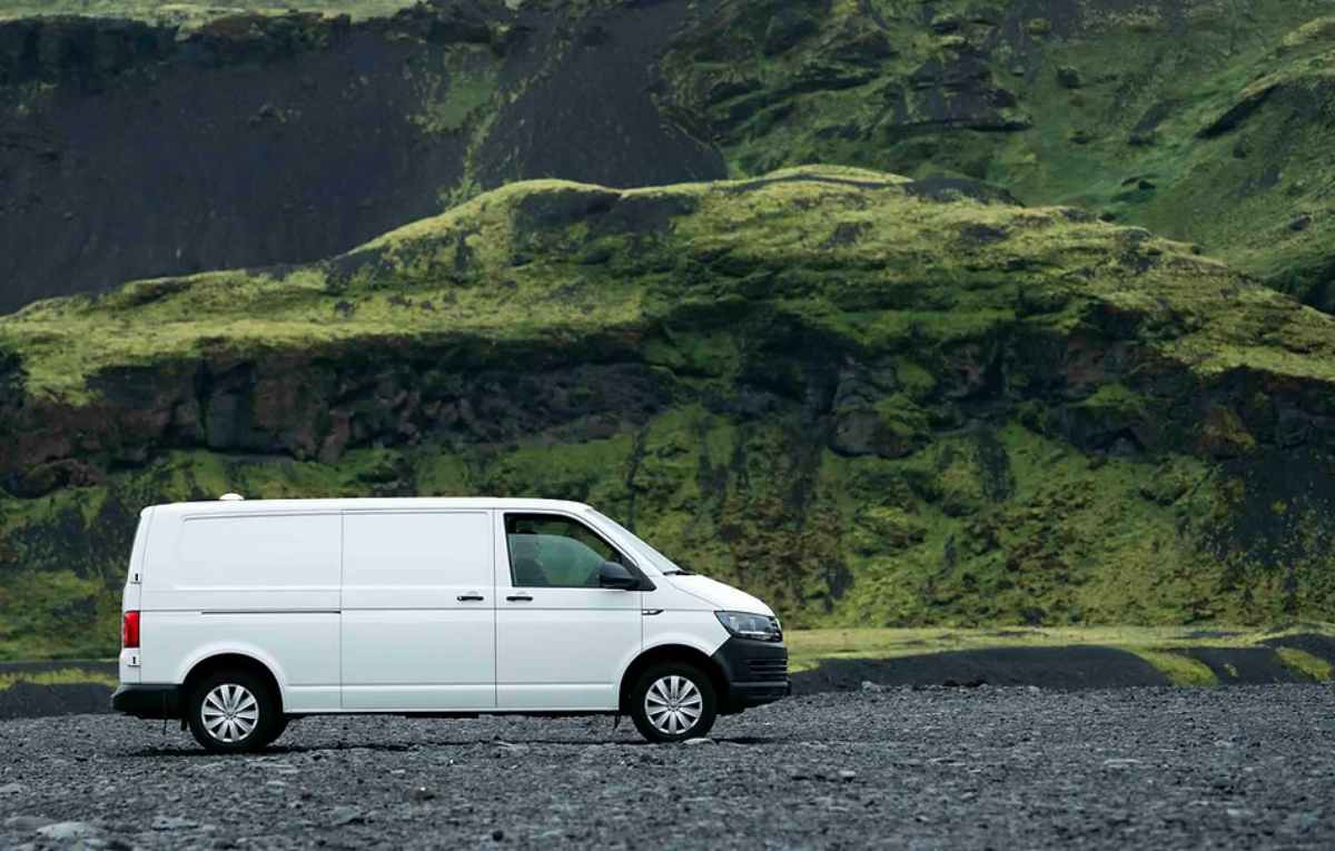 White VW Transporter parked on black volcanic plains with mossy cliffs rising in the background.