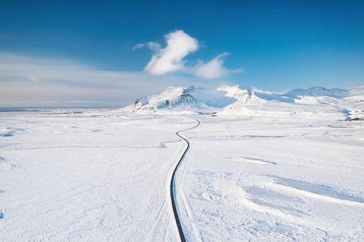 Aerial view of a lone paved road winding across a snow-blanketed plain toward jagged mountains under a bright blue sky in Iceland.