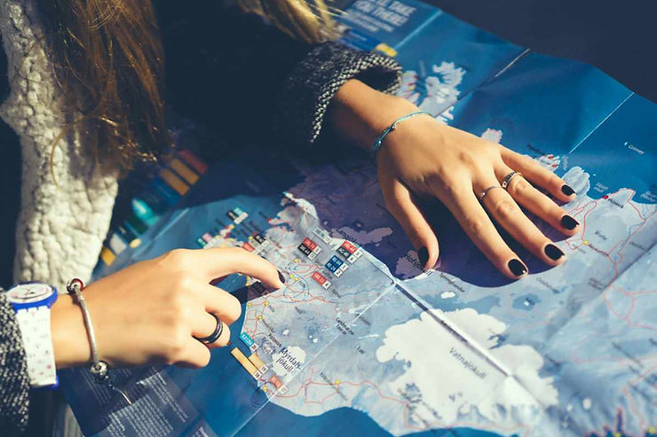 Traveler’s hands point to towns and icons on a detailed paper map of Iceland while planning a winter road trip route and stops.