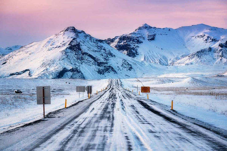Icy two-lane road leading toward snowcapped mountains at sunrise in Iceland, with warning signs, yellow posts, and a slick, windblown surface.