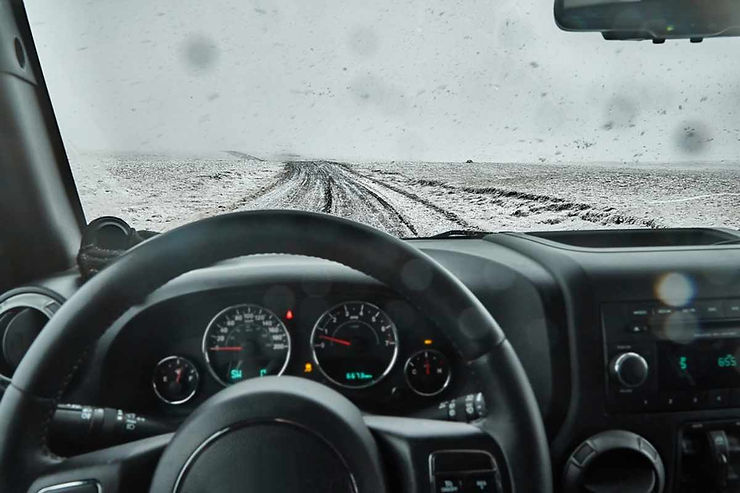 Driver’s view through a wet windshield toward a rutted, snow-covered track in Iceland; dashboard gauges visible, conditions muddy, slick, and remote.