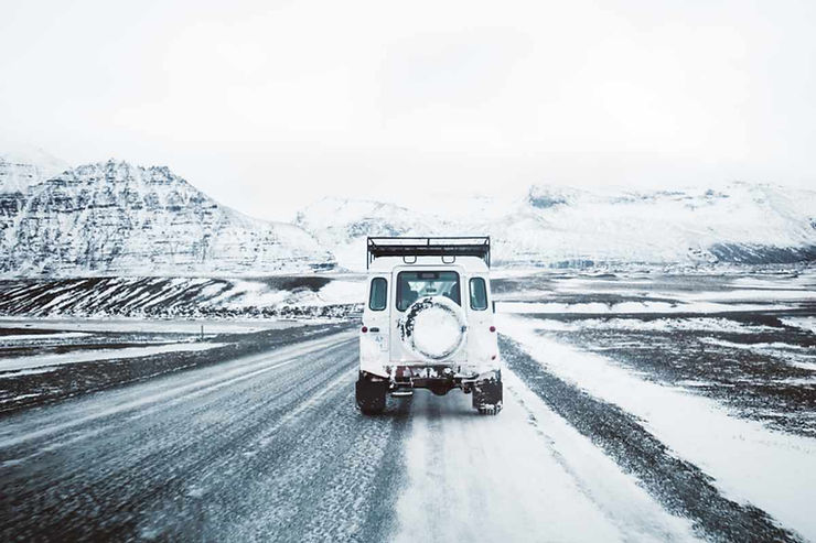 Rear view of a white 4x4 driving a snow-dusted gravel road through Iceland’s countryside, dark mountains ahead in flat, overcast winter light.
