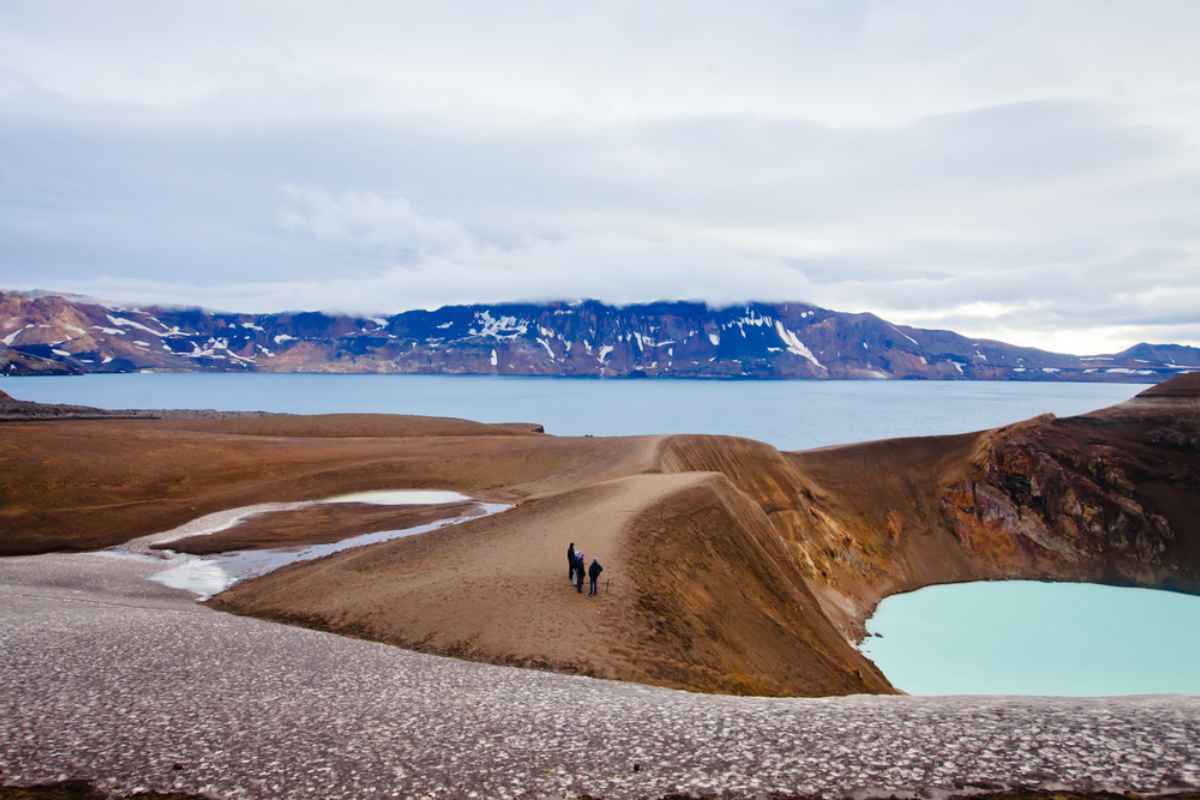 Hikers on a narrow ridge between a turquoise crater lake and barren slopes in Iceland’s Highlands, snowy peaks across the water.