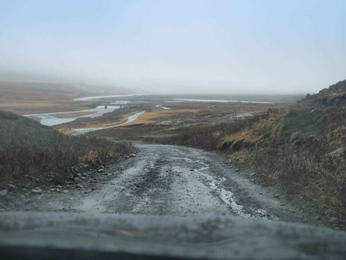 Muddy, rutted F-road through a misty Highland valley with braided rivers and a small bridge visible in the distance after rain.