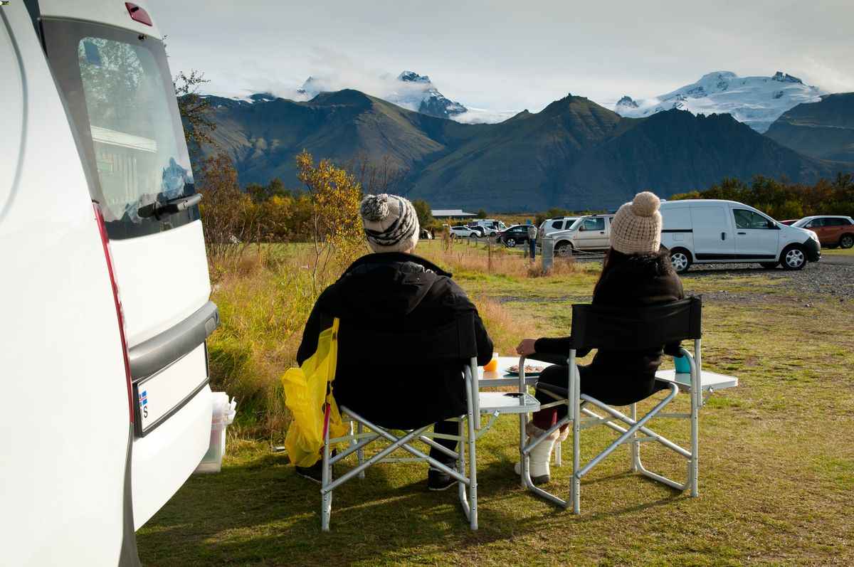 Couple camping beside a campervan in Iceland, eating outdoors with glacier-topped mountains in the background.