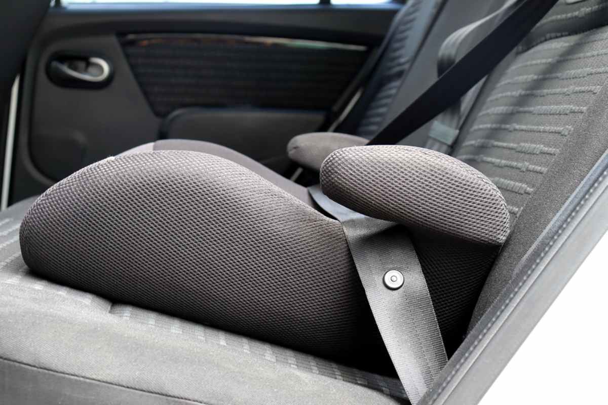 Black car seat with a child's booster seat strapped in, set against a patterned fabric interior. The seatbelt is fastened securely.