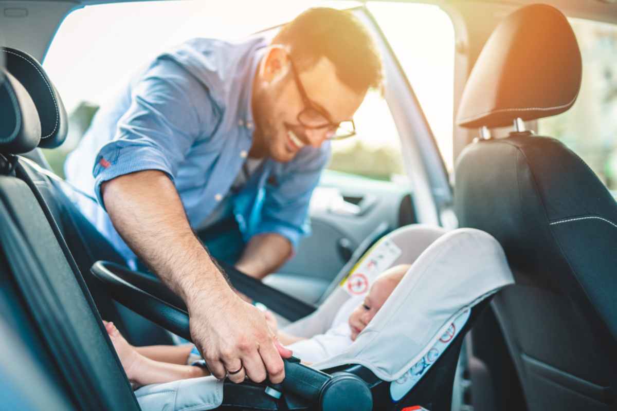 Man in blue shirt smiles while adjusting a baby's car seat inside a car. Sunlight streams in, creating a warm, caring atmosphere.