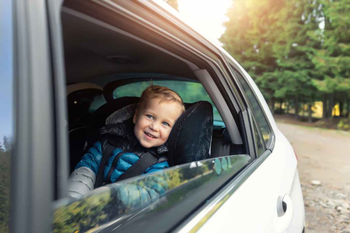 Child smiling from car window, wearing a blue jacket. Parked car on a forested road, sunlight filtering through trees. Playful mood.
