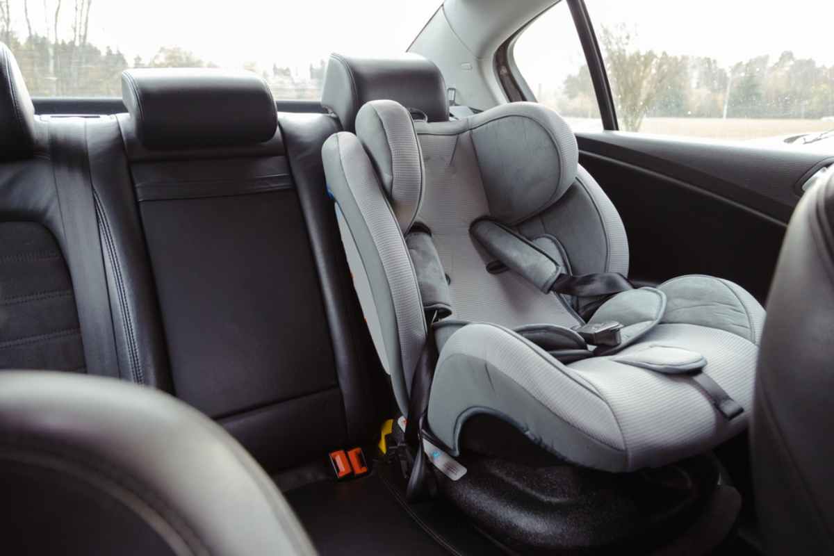 Gray car seat installed in a black car interior, rear seat. Background shows blurred outdoor scenery through windows.