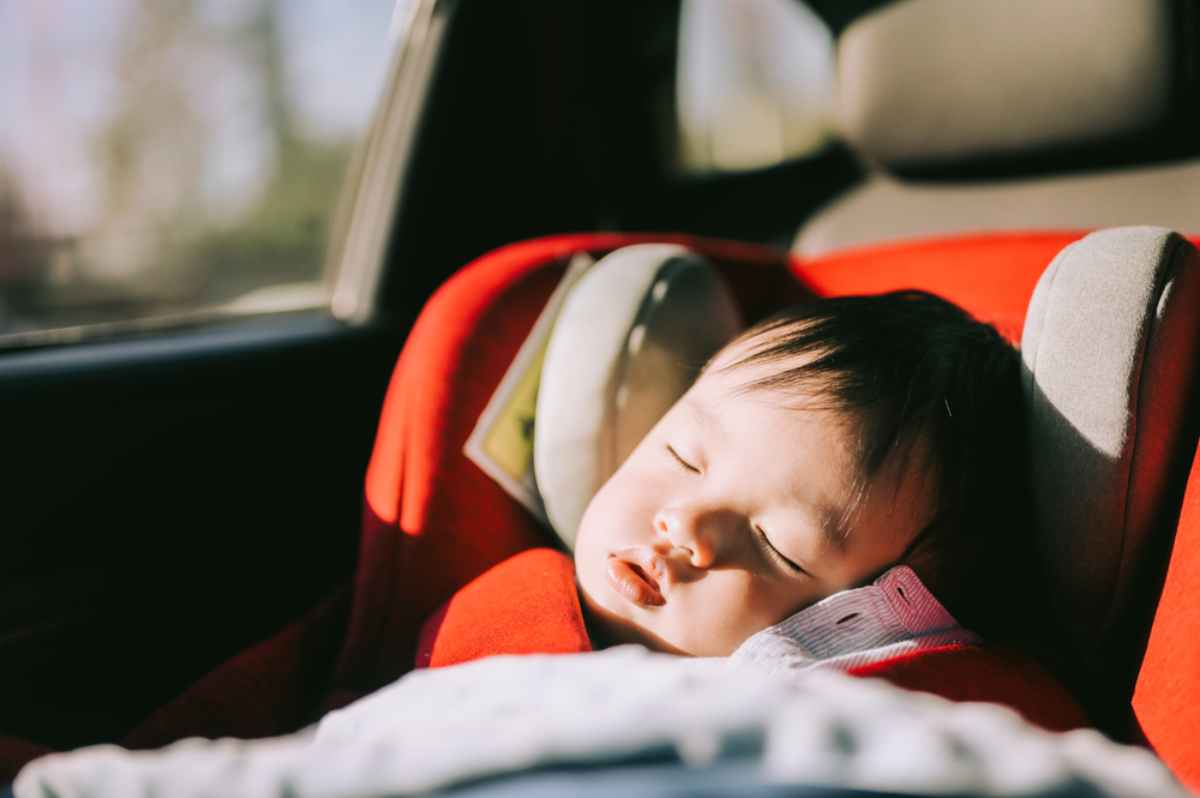 A baby sleeps peacefully in a red car seat with soft sunlight streaming in. The interior of a car is visible in the background.