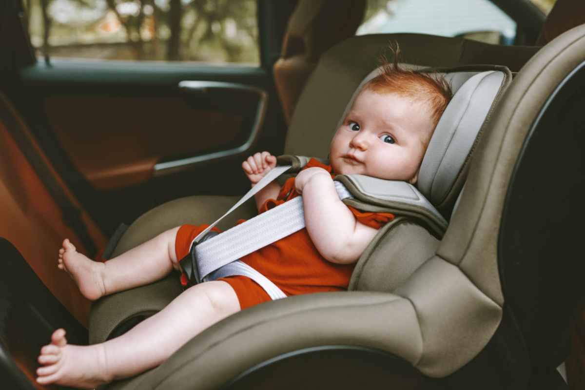 Baby in red outfit sits in a car seat, gazing quietly. Car interior visible, with soft light filtering through windows. Calm mood.