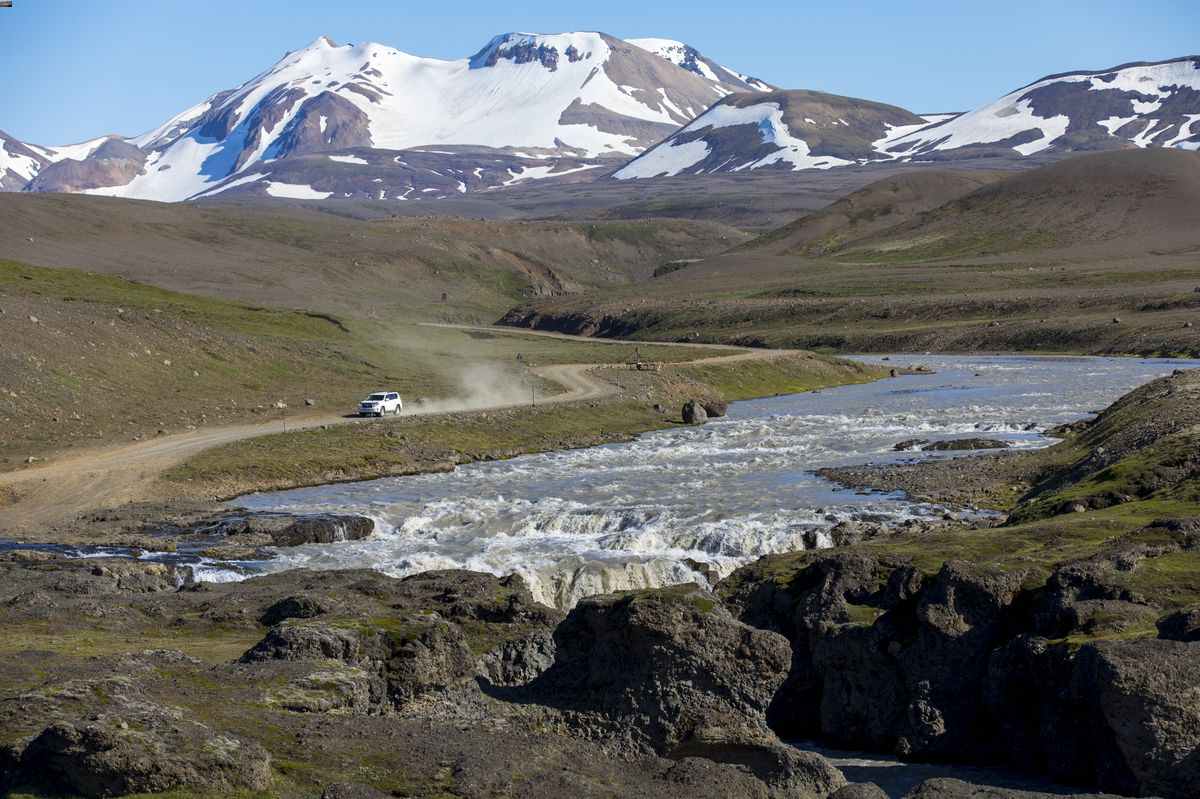 4x4 driving a gravel F-road beside a fast glacial river and snow-capped mountains.
